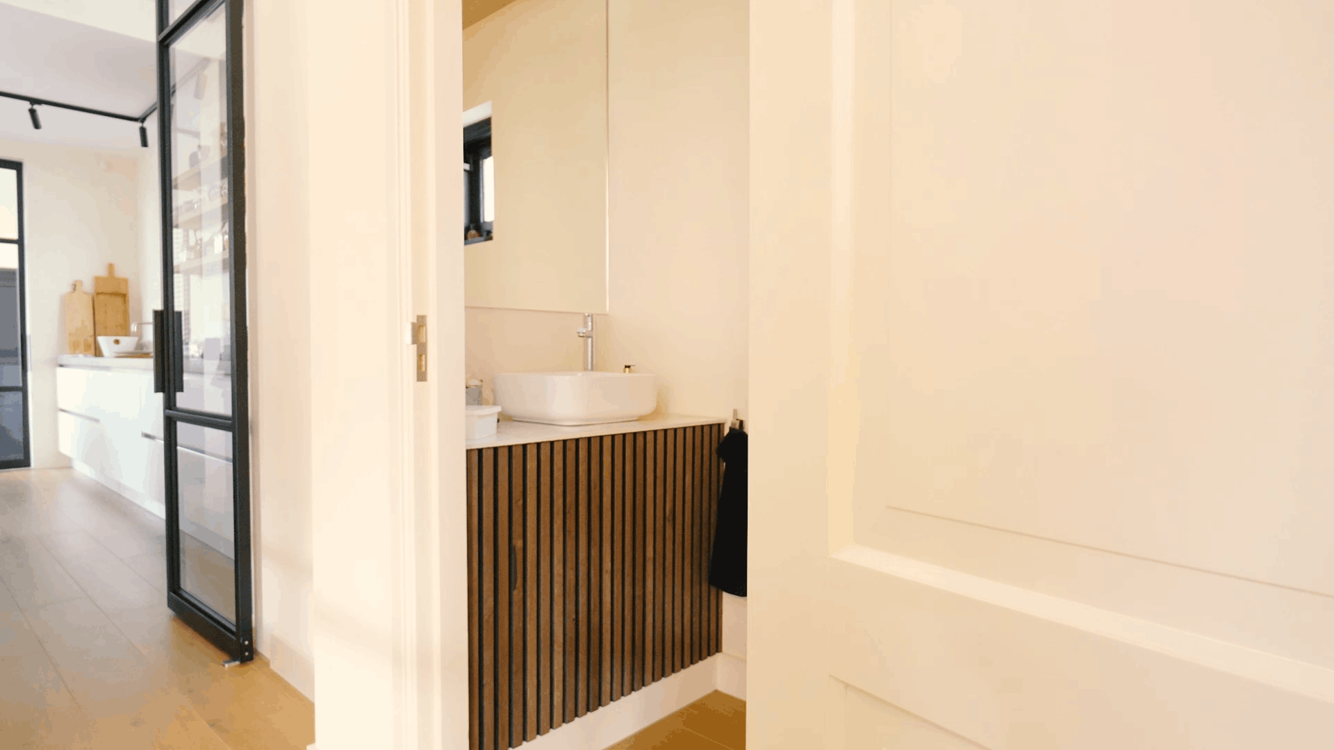 Modern bathroom featuring a round sink, wood slat acoustic vanity, large mirror, and white door next to a bright hallway and contemporary kitchen.